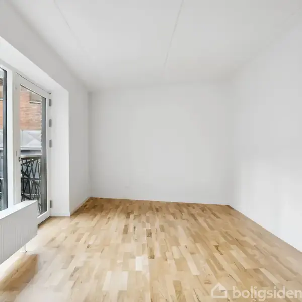 Tomt rum med trægulv og hvide vægge, hvor et stort vindue mod venstre giver udsigt til cykler udenfor. Lysindfaldet skaber et åbent og lyst miljø.