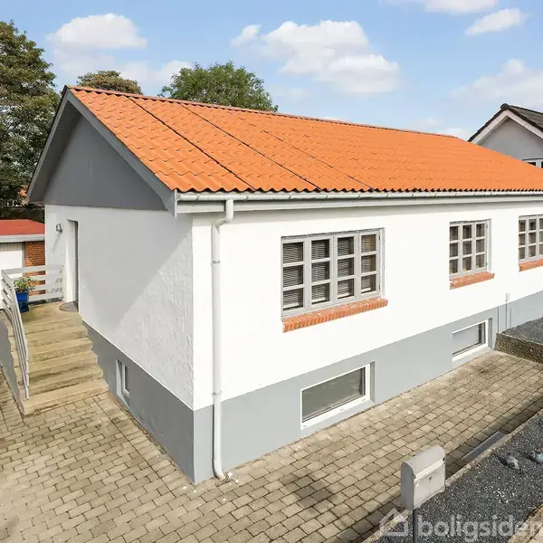 En hvid bungalow med rødt tegltag står på en flisebelagt indkørsel. Til venstre ses en garage. Baggrunden har træer, og en blå himmel med lette skyer skaber en rolig atmosfære.