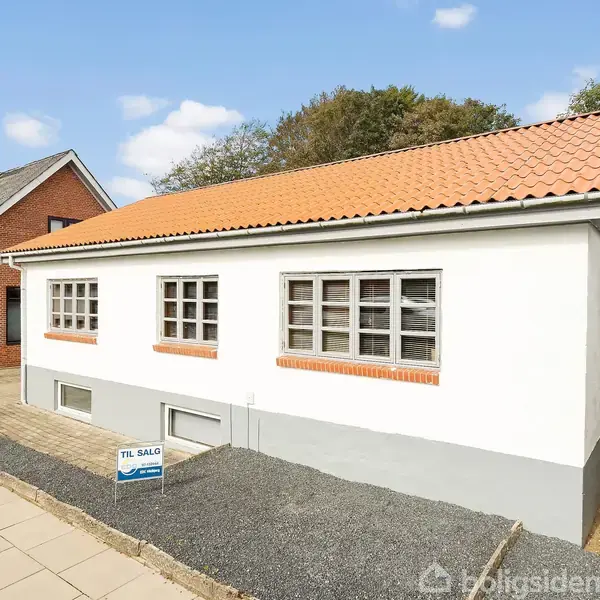Hvidt hus med rødt tegltag står til salg, ses fra gaden, omgivet af fortov og grusbelagt jord. Skiltet foran huset har teksten: "TIL SALG".