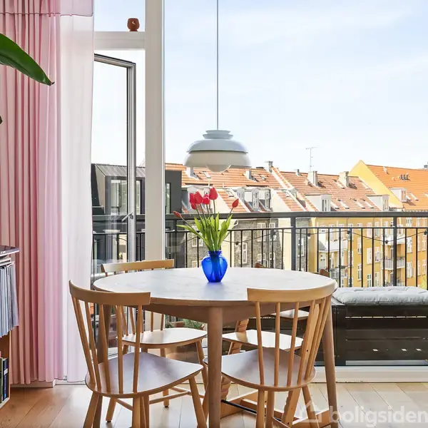 Rundt bord med vase og blomster står på trægulv i stue med åbne glasdøre mod altan. Bygninger ses i baggrunden under blå himmel. Reoler med bøger står til venstre.