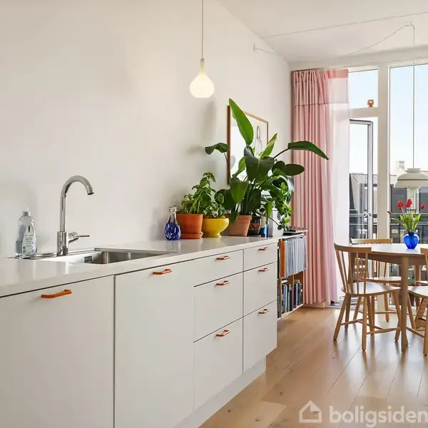 Køkken med hvide skabe og orange greb har en espressomaskine. Planter pynter i vindueskarmen. Bord og stole står ved et stort vindue med lyserøde gardiner, udsigt til bygninger udenfor.