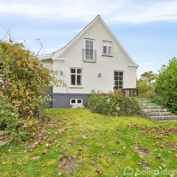 Et hvidt hus med saddeltag står stille i en have omkranset af buske og en stentrappe. Blade ligger spredt på det grønne græs.