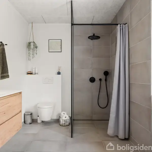 Badeværelse med en moderne brusekabine i glas og sort armatur. Ved siden af er et toilet og en trækommode med håndvask og spejl. Gråt håndklæde hænger på en krog.