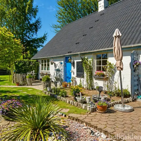Et hvidt hus med blå døre står i en frodig have. En markise er slået op, og der er blomster i sengene langs husets mur.