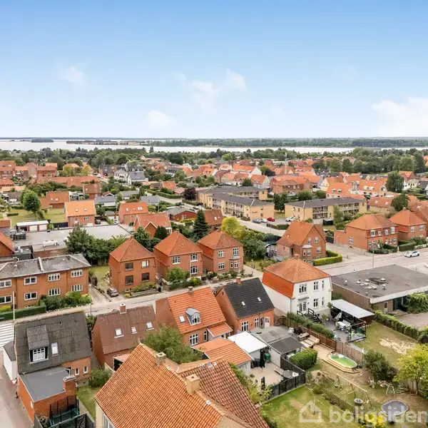 Røde murstenshuse med skrå tage står tæt i forstadskvarter, omgivet af veje og grønne haver, mens en fjernere skov og vandkant skaber naturlig baggrund.