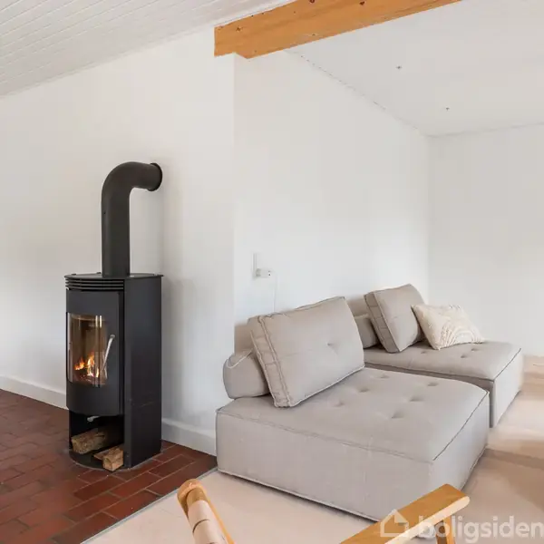 En moderne stue med en grå hjørnesofa, brændeovn tændt ved siden af, og lamper hængende fra loftet. Rummet har lyse vægge og træmøbler, hvilket giver en hyggelig atmosfære.