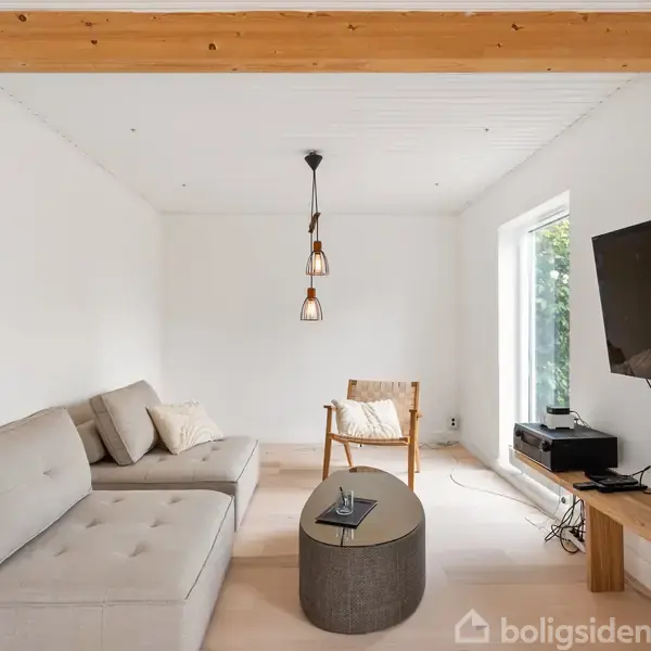 En stue med en grå sofa og en stol, begge vendt mod et vægmonteret TV. Et rundt bord står i midten. Indretningen er moderne med en hængende lampe.