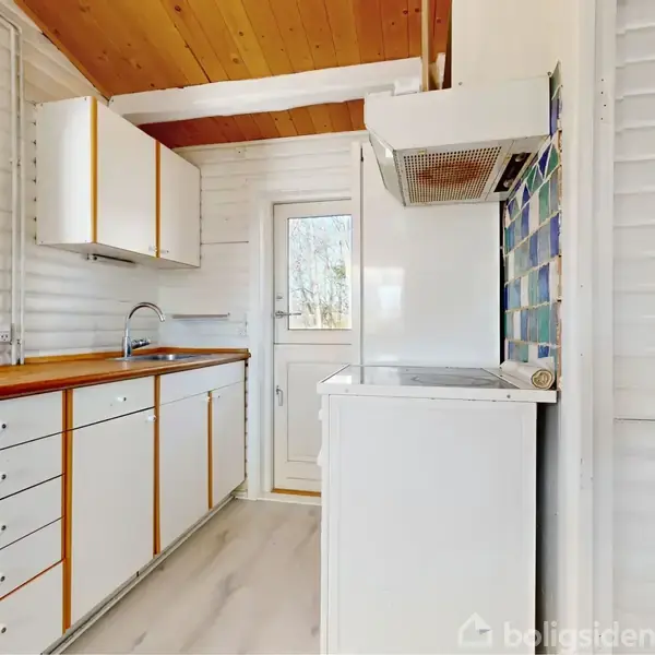Køkkenet består af hvide skabe og skuffer med trækanter, en træbordplade og komfur. En dør fører udenfor, og vinduer giver naturligt lys. Loftet er træbeklædt.