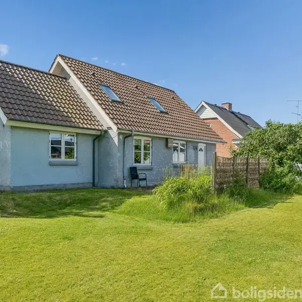 Et gråt hus med rødt tegltag står stille i en grøn have med græsplæne. Solrig dag, få skyer på himlen.