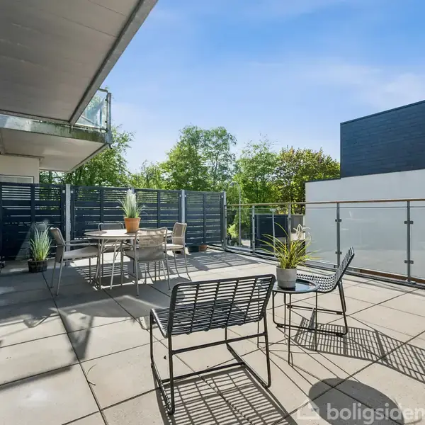 Terrasse med borde og stole, omgivet af glasrækværk. Flere potteplanter pynter området. Baggrunden viser moderne bygninger og grøn beplantning under en klar, blå himmel.