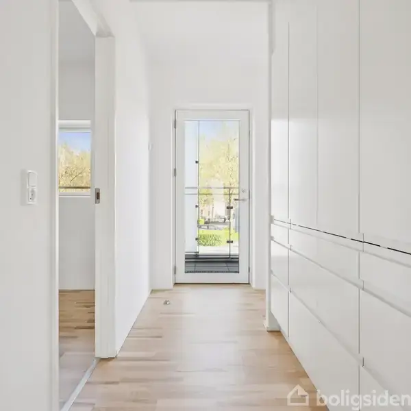 En lys gang forbinder værelser med hvidskabe på højre side og en glasdør for enden, der åbner ud til en balkon. Gulvet er lys trægulv.