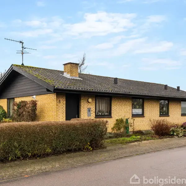 Et gult murstenshus står stille i et forstadsområde. Det har et sort tag, højre sidevindue og er omgivet af buske og træer langs en stille vej.