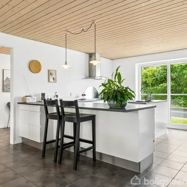Køkken-ø med grøn plante i moderne køkken. To sorte barstole ved disken. Store vinduer bagved giver udsigt til grøn have. Træloft og sorte fliser skaber elegant atmosfære.
