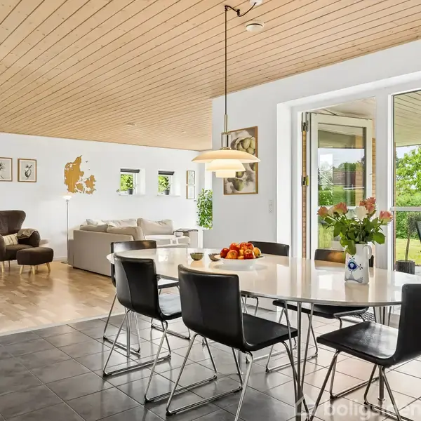 Et spisebord med sorte stole står ved siden af en vase med blomster og en skål med frugt. I baggrunden er en stue med hvide møbler og store vinduer, der åbner ud til en terrasse med grøn udsigt.