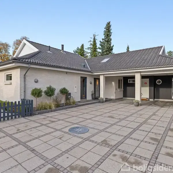 Et moderne, enkelt etplanshus med gråt tegltag står på en rummelig flisebelagt gårdsplads omgivet af grønne træer og buske, der skaber en harmonisk og fredelig atmosfære.