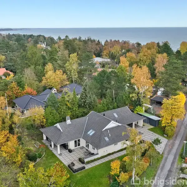 Hus med gråt tag ligger ved en snoet vej, omgivet af efterårsfarvede træer. Flere huse og en skov fylder landskabet, og havet ses i baggrunden.