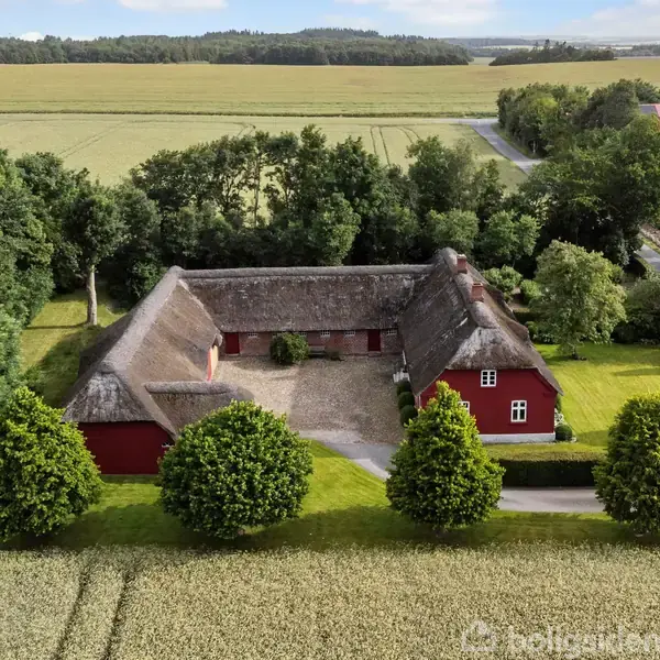 En stor, rød gård med stråtag danner en firkantet struktur i en frodig landzone omgivet af træer og marker. En indre gårdsplads er synlig mellem bygningerne.