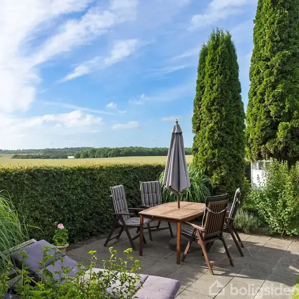 Terrassebord med parasol og stole står på en flisebelagt gårdhave. Omkring er der frodig bevoksning og høje tujaer, som indrammer det åbne landskab i baggrunden under en klar blå himmel.