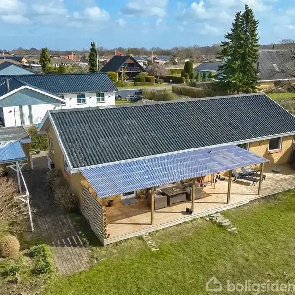 Et hus med mørkt tag står med en overdækket terrasse i en have. Flere huse ligger tæt på, og himlen er let overskyet med spredte skyer.