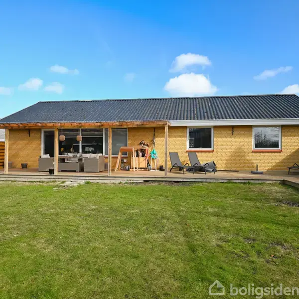 En gul murstenshus med sort tag står i solrigt vejr. Møbler og liggestole er placeret på en træterrasse. Græsplæne omgiver huset, og himlen er klar og blå.