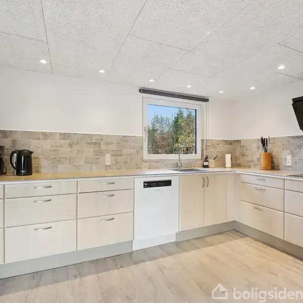 Et moderne køkken med lyse træskabe og skuffer, står i en rummelig indretning. En opvaskemaskine og køkkenredskaber er på bordpladen under et vindue med udsigt til træer.