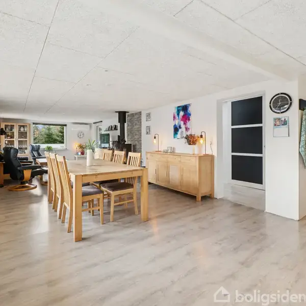 En spisestue med et langt træbord og otte stole står i et lyst, rummeligt rum. Til venstre ses store vinduer med planter, og til højre er et køkkenområde.