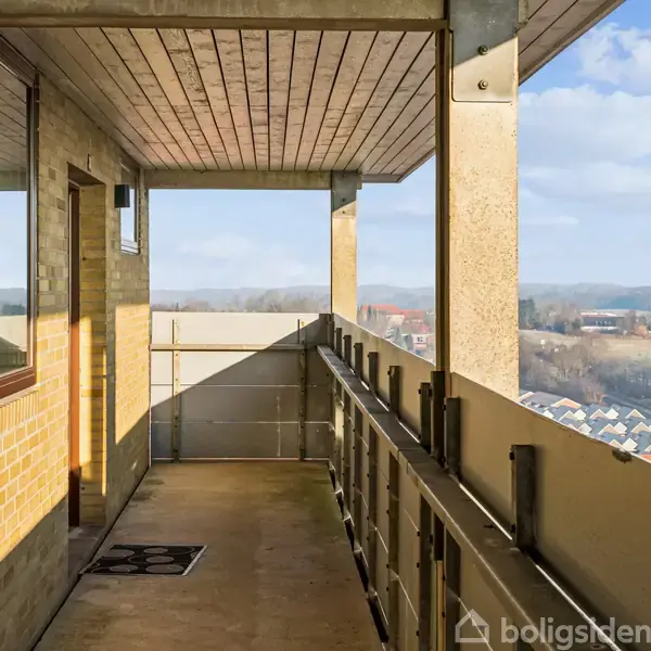 Balkon med gelænder, vender mod udsigt over hustage i et forstadsområde. Sollys skinner på gul og murstensvæg. Linoleumstæppe er placeret nær en dør til venstre.