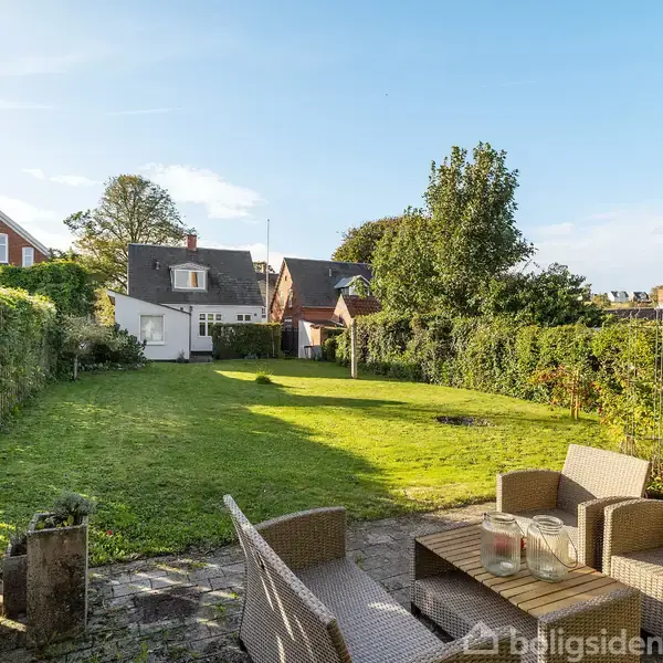 Havemøbler står på en terrasse, omgivet af en velplejet græsplæne og grønne hække, med huse og træer i baggrunden under en klar himmel.