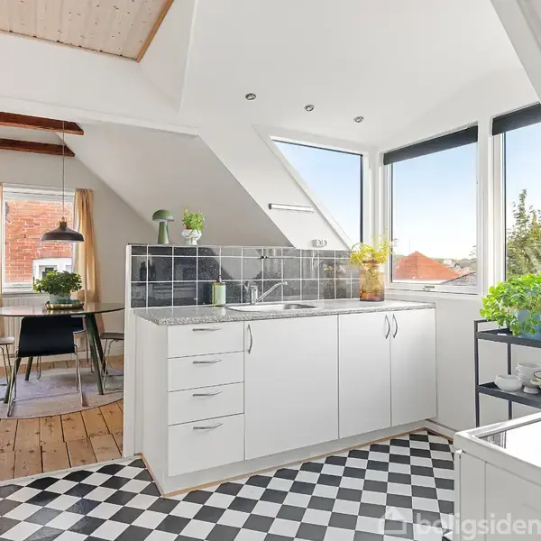 Hvidt køkken med sort-hvidternet gulv, hvor blomster og planter står på bordpladen. I baggrunden er der et spisebord ved et skråvinduet med udsigt over rødt tag og træer.