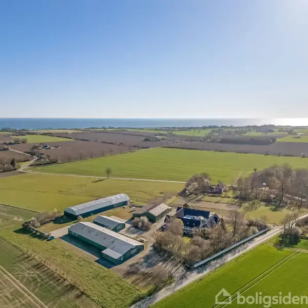 Gårde og bygninger ligger spredt ud over landbrugsjord. I baggrunden ses skovområder og marker under en klar blå himmel med havet i horisonten.