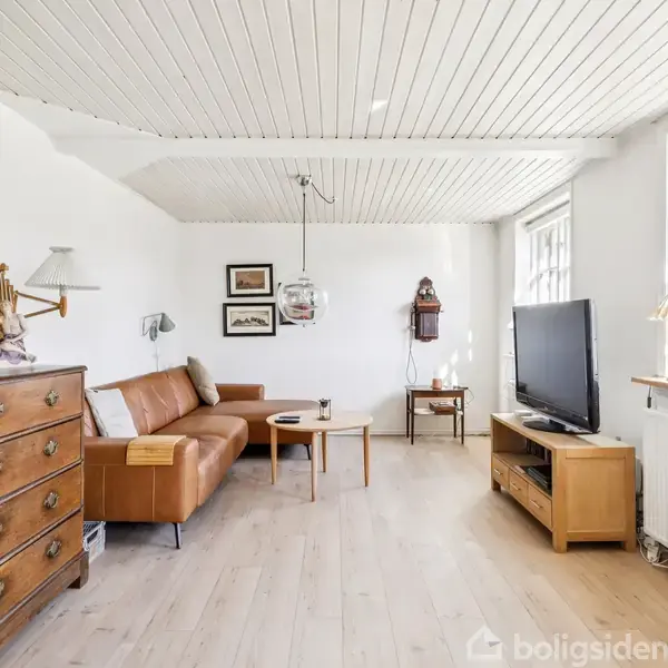 Brun lædersofa står mod væggen i en lys stue. Sofaen er omgivet af et træbord og tv-møbel. Væggene er dekoreret med spejle og billeder.