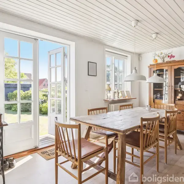Spisebord med seks stole står i et lyst rum med trægulv og hvidt træloft. En åben dør fører til en have. Skab og lampe er i baggrunden.