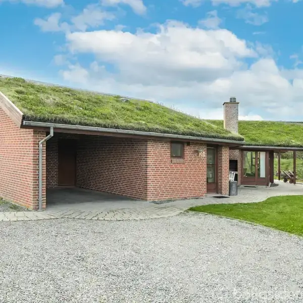 Rødt murstenshus med græstag står på en grusbelagt grund. Ved siden af er en overdækket terrasse og en carport. Huset er omgivet af grønne marker og under en blå skyet himmel.