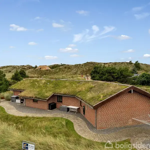 Hus med græstag står stille i et kuperet landskab med sandklitter og spredte træer. Murstensfacaden suppleres af en stenbelagt sti, der omkranser bygningen.