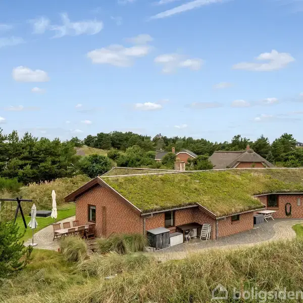 Et rødt murstenshus med græstag står stille omgivet af naturlige klitter og grønne buske. En terrasse med parasol og gyngestativ er synlig i baghaven under en blå himmel med skyer.