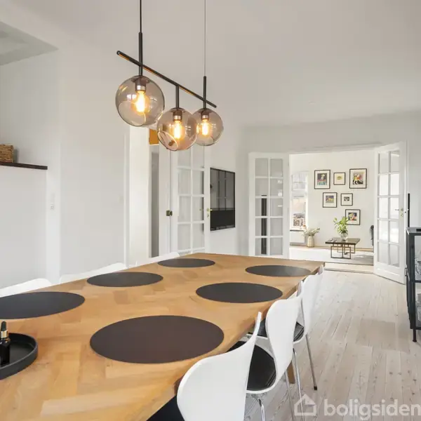 Spisebord i træ med sorte dækkeservietter, omgivet af hvide stole, i et lyst rum. Hængelampe med glødepærer over bordet. I baggrunden ses en opholdsstue med kunst på væggene.