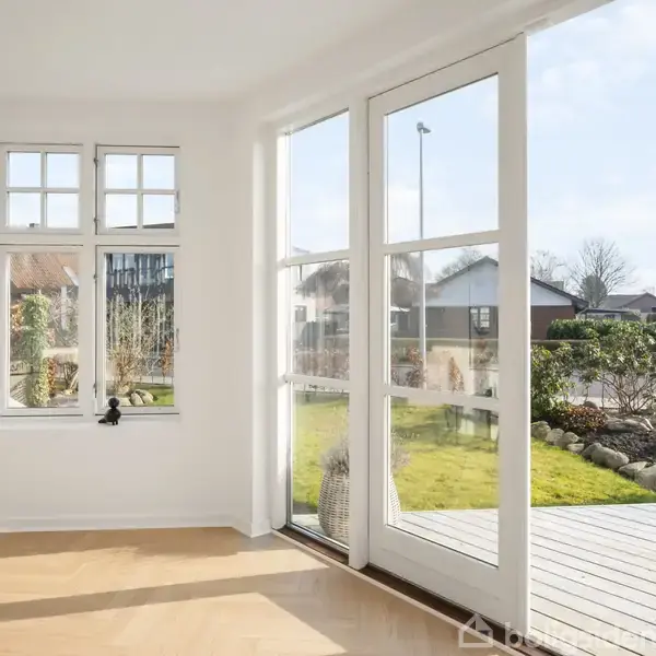 Lyse glasdøre åbner mod en solrig terrasse med en grøn have. Indendørs trægulve og store vinduer skaber et lyst rum. Udsigt til en stille villavej og nabohuse.