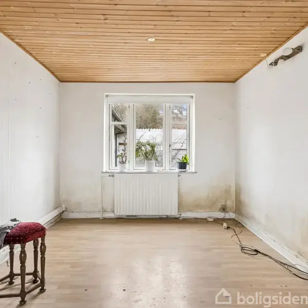 Stol står ved væg; tomt rum med træloft og stort vindue. Gavlvægge er slidte med fugtpletter. Lampe hænger på væggen. Gulvet er lyst, og en reol står i hjørnet.