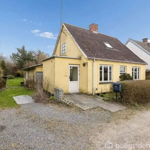 Et gult hus med en skråt tag står ved siden af en grusvej. Foran huset er en lille terrasse og en blå postkasse. Hækken adskiller haven fra vejen.