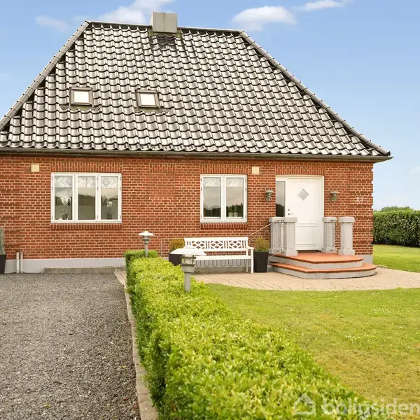 Et rødt murstenshus står stille med mørkt tegltag, omgivet af en velplejet have og en grusbelagt indkørsel. En hvid bænk ses ved hoveddøren.