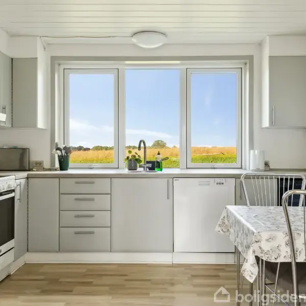 Et spisebord står i et rum med stenvæg, omgivet af seks brune stole og en brændeovn. Vinduerne til højre har et gardin, og der er potteplanter på vindueskarmen.