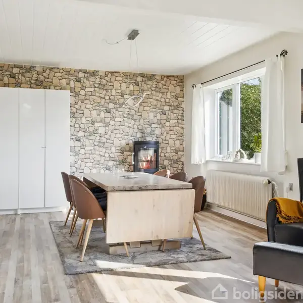 Spisebord omgivet af stole står på et tæppe i et rum med stenvæg og brændeovn. Et stort vindue lyser rummet op, og et billede af en løve hænger på væggen.