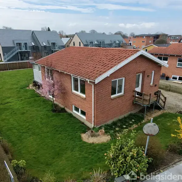 Et rødt murstenshus med skråt tag står på en græsplæne. Blomstrende buske omgiver huset. I baggrunden ses flere boliger, og en vej løber langs siden.