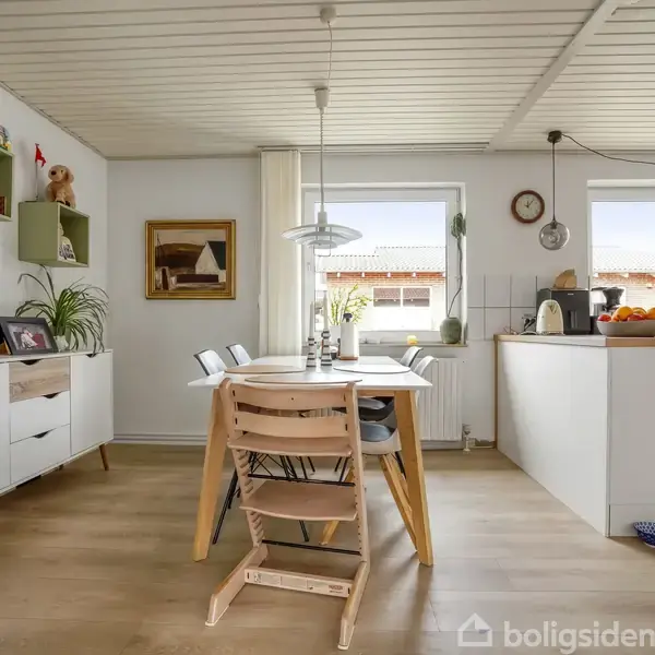 Et lyst køkken-alrum med et rundt spisebord og træstole, omgivet af hvide vægge, reoler med planter og vinduer med udsigt til omgivelserne. Der hænger en lampe over bordet.