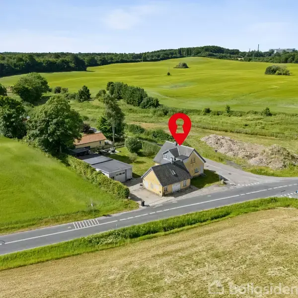 Et gult hus står ved en landevej, omgivet af grønne marker og skovområder. Et rødt nåleikon markerer huset. Landskabet er åbent og landligt.