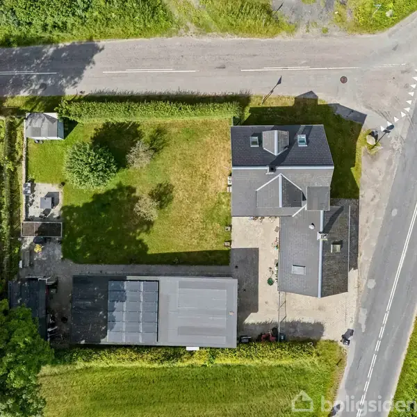 Hus med mørkt tag ses ovenfra, omgivet af en stor grøn have. I nærheden er der en garagebygning og en lang vej på højre side.