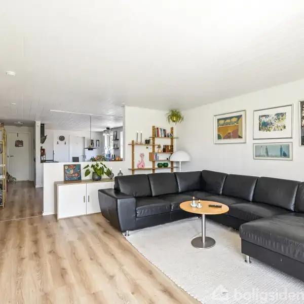 Sort lædersofa står i en stue med trægulv. Væggen har flere malerier. Et rundt bord står foran sofaen, og et TV er placeret modsat. Køkken er synligt i baggrunden.