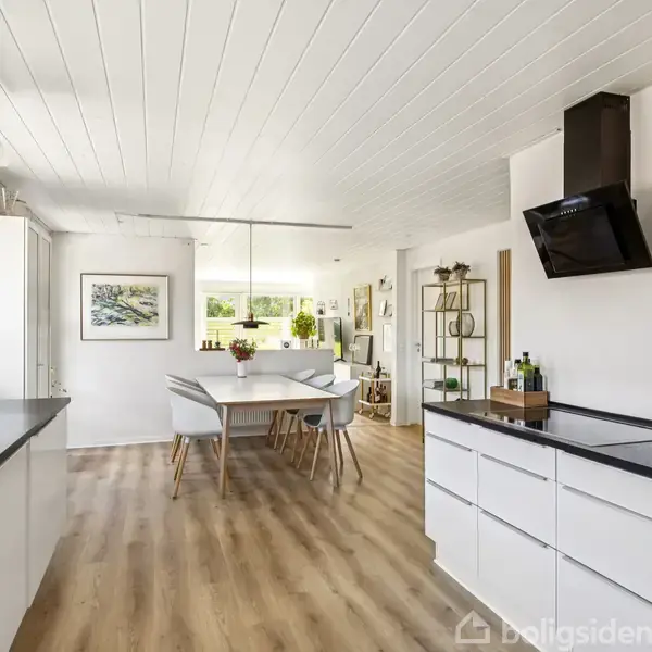 Køkken med hvide skabe og sort bordplade. Et spisebord med stole står i midten. Store vinduer giver naturligt lys. Trægulv og hvidmalet loft skaber en lys atmosfære.