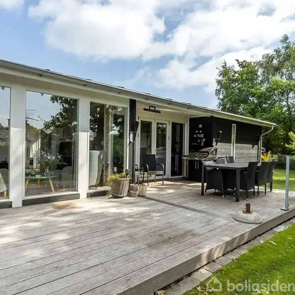 En terrasse med havemøbler og grill står foran et moderne hus med store vinduer. Terrassen er omgivet af en grøn have under en blå himmel med skyer.