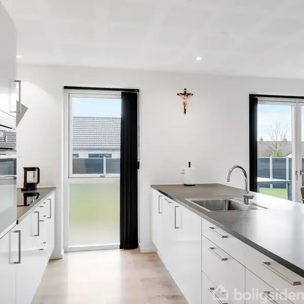 Køkken med hvidt interiør og sort bordplade, set i daglys. På væggen hænger et krucifiks. Store vinduer giver udsigt til husets have. En ovn er indbygget i skabet.